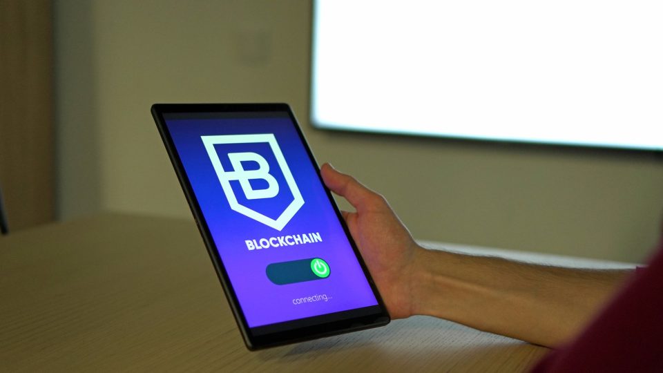 Pessoa segurando um tablet com um símbolo de blockchain na tela, representando tecnologia de redes digitais.