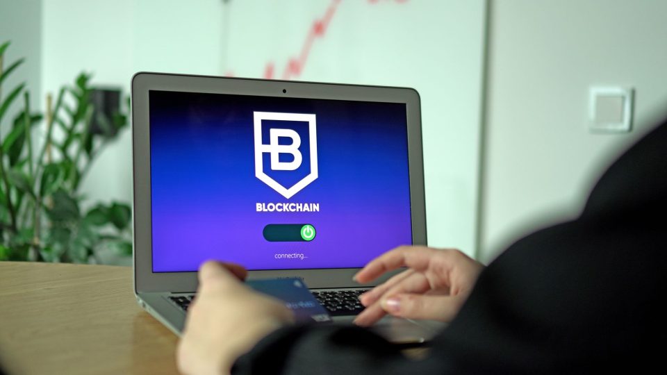 Mão segura um cartão diante de um laptop que exibe uma tela de conexão com blockchain.